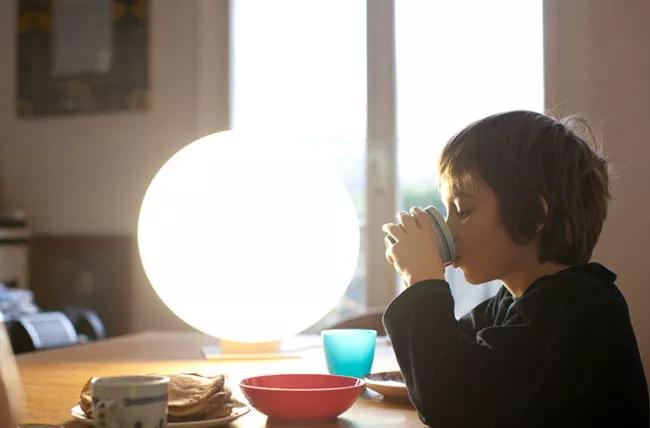 Bambino seduto a un tavolo accanto a una lampada, mangiando e bevendo.